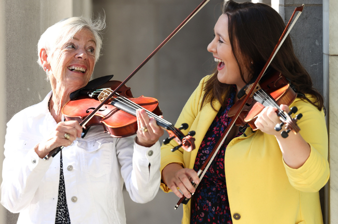 Two women play the fiddle together in the sunshine