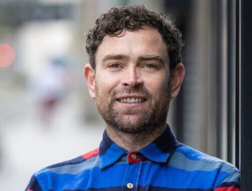 A man with short curly hair and a beard wearing a colourful patterned shirt is leaning against a wall with his arms crossed over his chest in a street