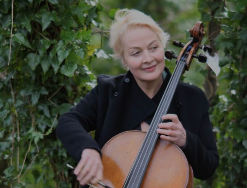 A woman with short blonde hair wearing a black jacket is playing a cello in a graden surrounded by green bushes