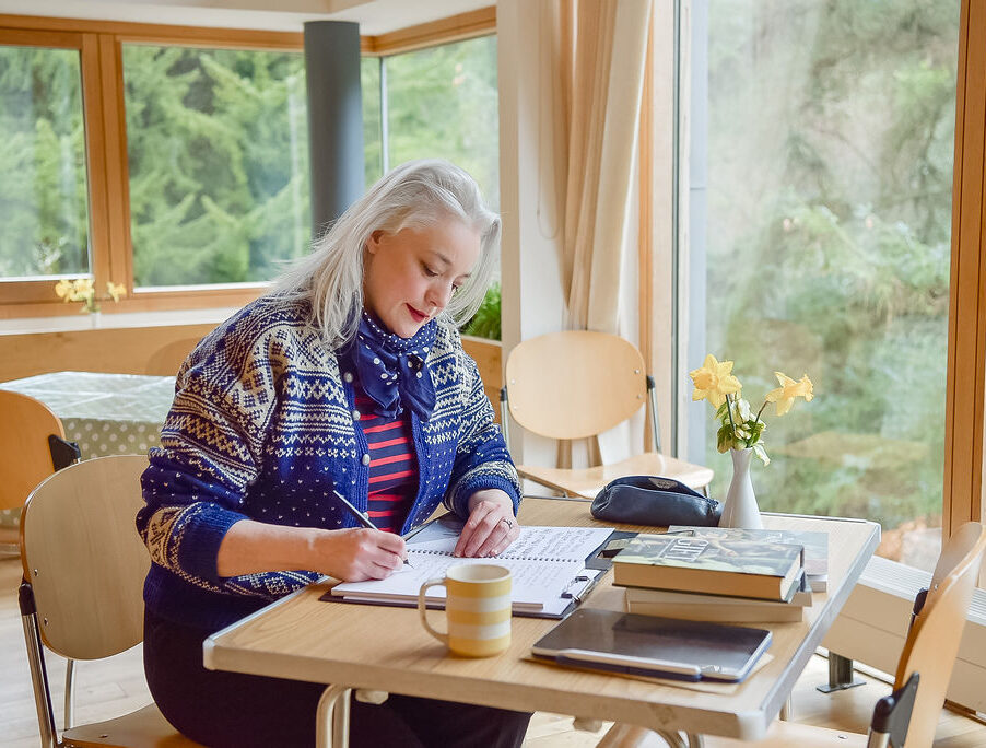 A women with long blonde hairs sits at a desk writing in a notebook in the background are windows with views of the trees outside