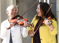 Two women play the fiddle together in the sunshine