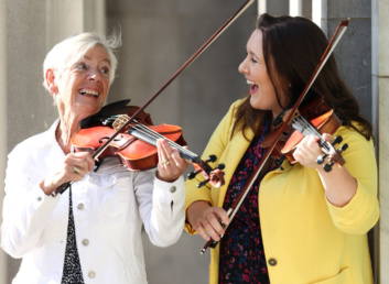 Two women play the fiddle together in the sunshine
