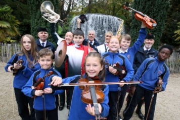 A group of children and adults dressed in school and band uniforms stand in front of a waterfall holding their musical instruments in the air